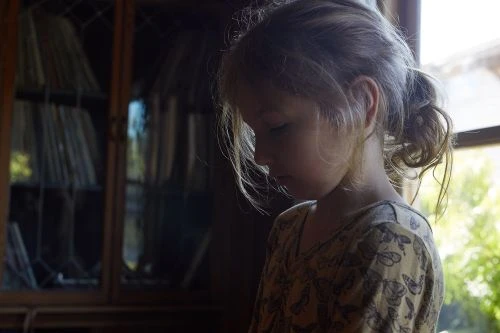 Profile portrait of a young child with a ponytail wearing a patterned top indoors near a wooden cabinet.