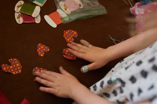 Child's hands working with heart-shaped fabric pieces on a brown surface with sewing supplies, creating a crafting project.