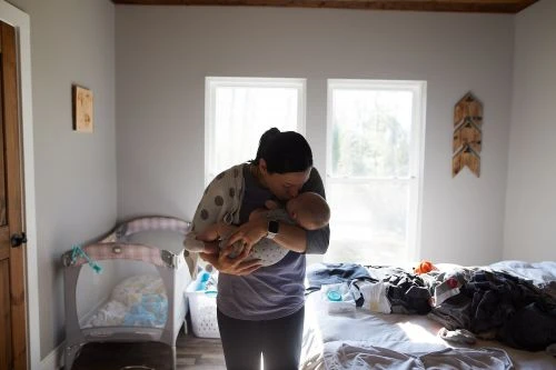 Woman in grey shirt and leggings holds a baby in indoor bedroom setting with daylight shining through windows, crib, and unmade bed.