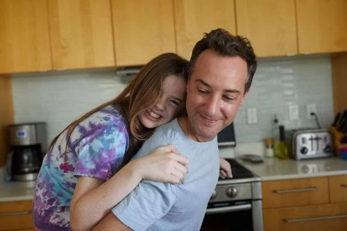A smiling father gives his daughter a piggyback ride in a bright kitchen with wood cabinets, stainless steel appliances, and white backsplash.
