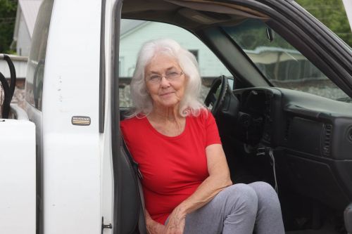 Elderly woman sits inside a white pickup truck wearing red t-shirt and grey sweatpants looking directly at camera with neutral facial expression.