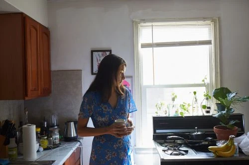 Woman in a blue floral dress standing in a kitchen holding a mug by a window with houseplants