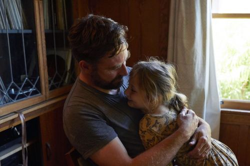 A bearded man embraces a young girl in a softly lit indoor space near a window and a dark wooden cabinet.