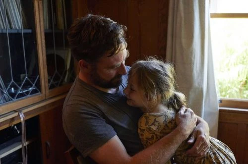 A bearded man embraces a young girl in a softly lit indoor space near a window and a dark wooden cabinet.