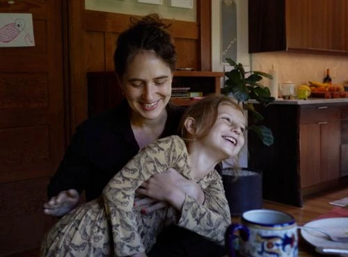 A smiling mother hugs her laughing young daughter in a bright indoor kitchen scene with hardwood floors and a houseplant in the background.