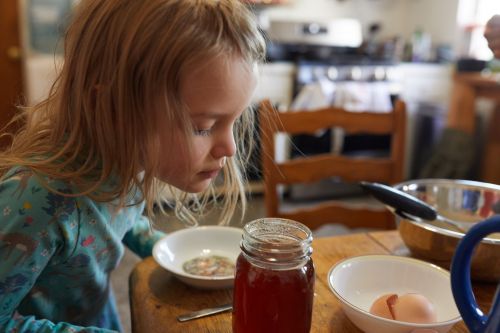 Young girl with blonde hair looks at a bowl on a wooden table in a kitchen setting. There is a jar of liquid and egg shells nearby.