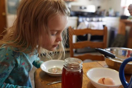 Young girl with blonde hair looks at a bowl on a wooden table in a kitchen setting. There is a jar of liquid and egg shells nearby.