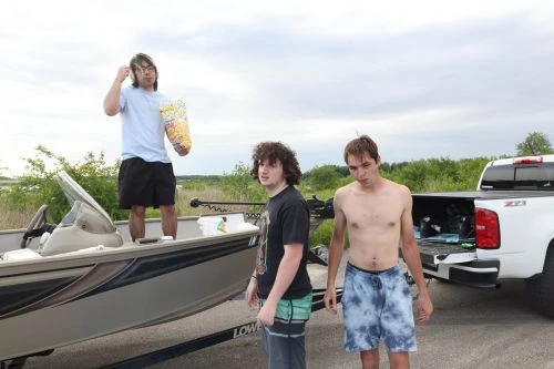 Three young men outdoors with a boat and truck under a cloudy sky; one holds popcorn, another is shirtless, capturing a casual moment.