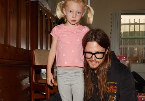 A blonde child with pigtails poses next to an adult male with long hair and glasses, indoors in front of a dark wood cabinet.