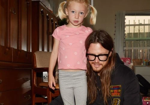 A blonde child with pigtails poses next to an adult male with long hair and glasses, indoors in front of a dark wood cabinet.