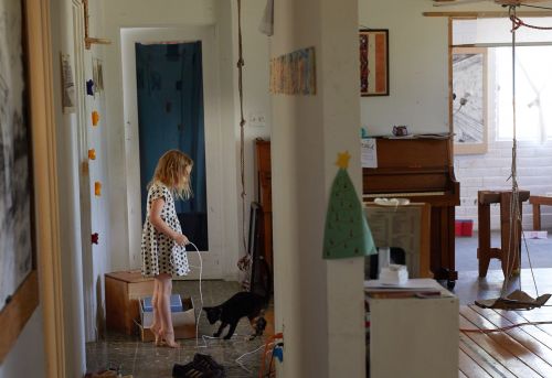 A girl with blonde hair in a polka-dot dress stands near a black cat inside a home with a piano and art, creating a candid indoor scene.