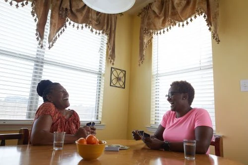 Two adult women joyfully play cards at a table indoors, illuminated by natural light streaming through window blinds, alongside fruit and drinks.