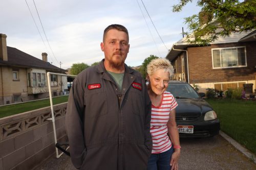 A man and woman are posing outdoors in front of houses and a car; the man is wearing a work uniform, and the woman is wearing a striped shirt.