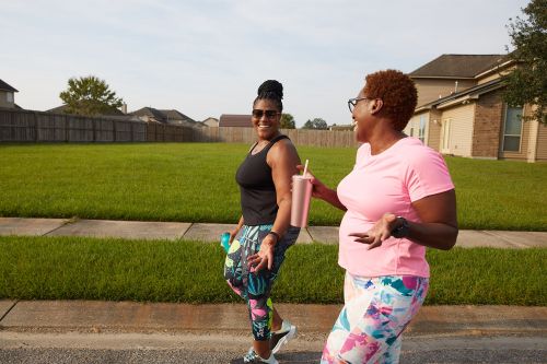 Two black women walking along a residential sidewalk, talking and smiling while holding reusable cups.