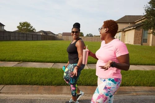 Two black women walking along a residential sidewalk, talking and smiling while holding reusable cups.