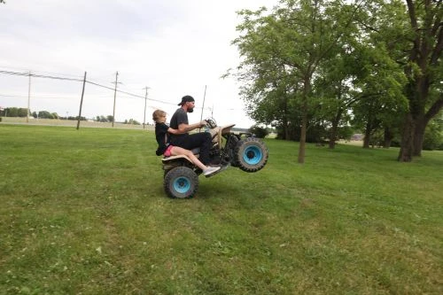 An adult and child are on a vintage ATV with one wheel raised on a grassy lawn with scattered trees under a cloudy sky.