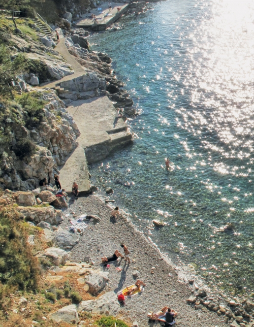 Scenic coastal view of people relaxing on a pebble beach by clear blue water, with rocky cliffs and sunlit water, ideal for summer vacation.
