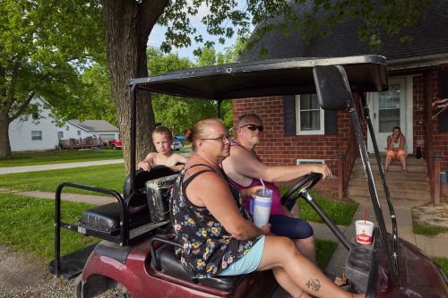 Adults and child in a red golf cart on a sunny street with houses and green trees in the background, enjoying an outdoor leisure activity.