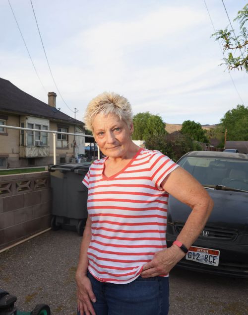 Adult woman poses outdoors in casual clothing with a house, car, and yard visible during daylight hours.