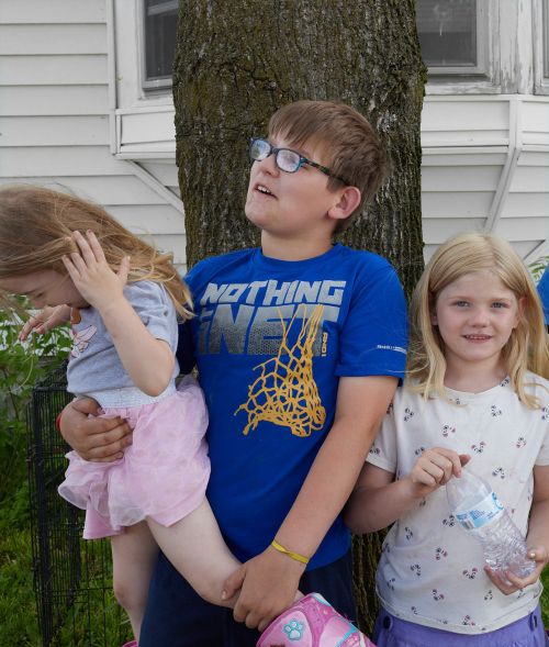 Portrait of three children outdoors by a large tree, featuring a boy wearing glasses and a blue graphic t-shirt holding a younger child.