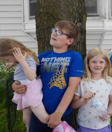 Portrait of three children outdoors by a large tree, featuring a boy wearing glasses and a blue graphic t-shirt holding a younger child.