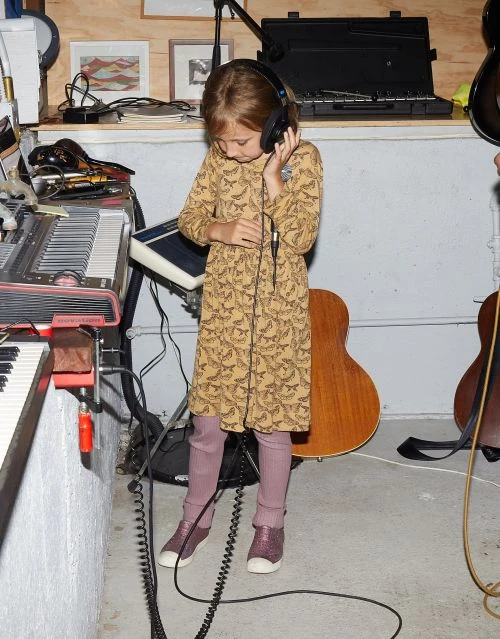 Young girl wearing headphones, a patterned dress, and mauve leggings, standing near musical instruments in an indoor space with concrete floors.