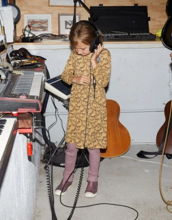 Young girl wearing headphones, a patterned dress, and mauve leggings, standing near musical instruments in an indoor space with concrete floors.