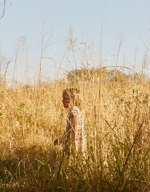 A young child in a patterned dress walks through a field of tall, golden grasses under a clear blue sky, framed by stalks of wheat.