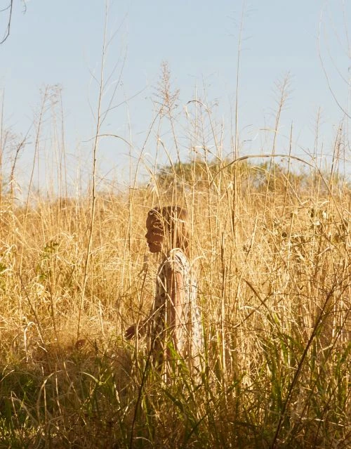 A young child in a patterned dress walks through a field of tall, golden grasses under a clear blue sky, framed by stalks of wheat.