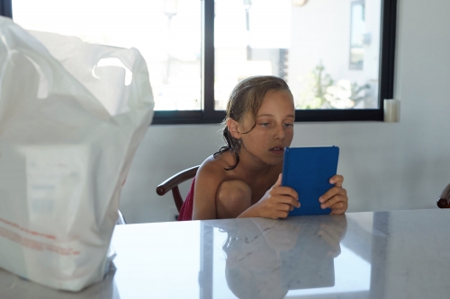 A child is seated at a table inside a room, holding a blue tablet, with a large white bag in the foreground and a window behind.