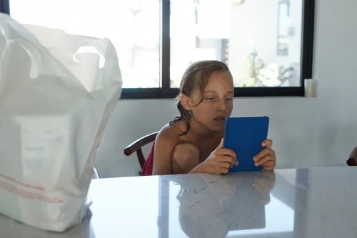 A child is seated at a table inside a room, holding a blue tablet, with a large white bag in the foreground and a window behind.