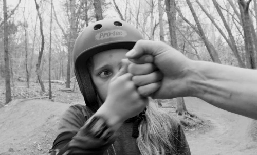 Black and white shot of a child wearing a helmet fist-bumping another person in an outdoor wooded area. The child is in focus.
