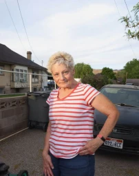 Elderly woman with short blonde hair wearing a red and white striped t-shirt posing outside next to a car