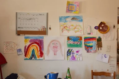 White wall covered with various children's drawings, paintings, and a dry-erase calendar board photographed by Douglas Adesko.