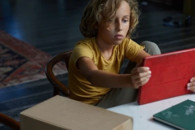 Young child with curly hair wearing a mustard yellow t-shirt seated at a table holding a red tablet