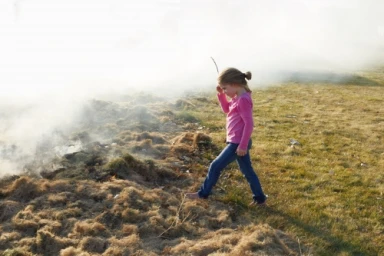 Young girl in pink top and blue jeans walking through smoke and dry grass outdoors photographed by Douglas Adesko.