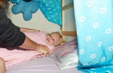 Child lying in bed with pink blanket while an adult tucks them in under blue patterned curtains