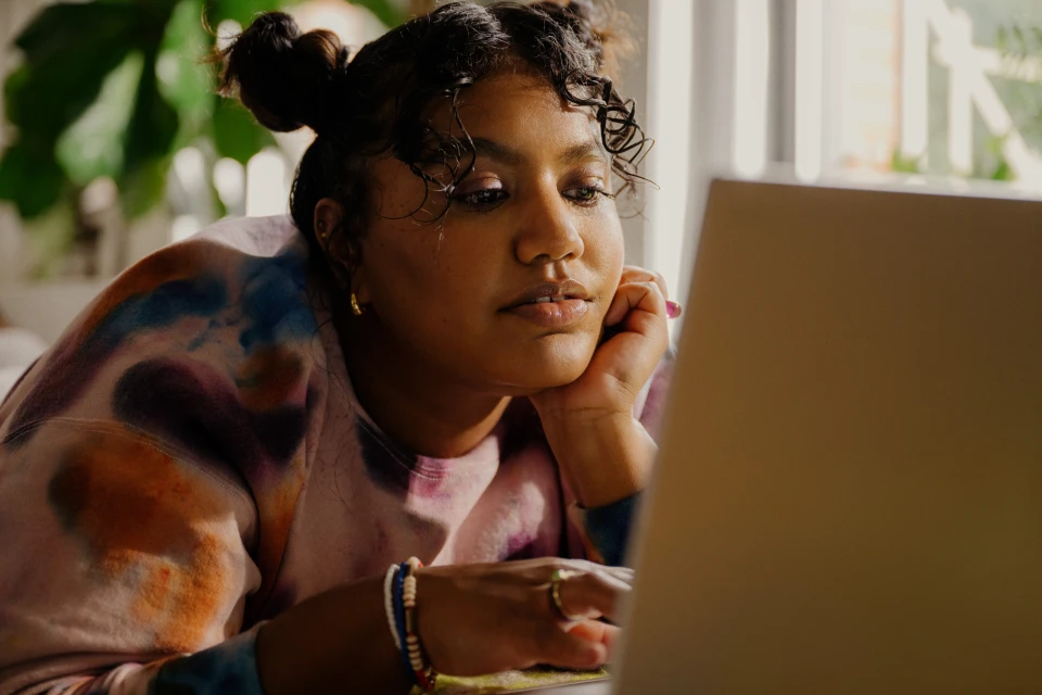 Young adult female with curly hair, wearing a tie-dye sweatshirt, looking at a laptop in a naturally lit indoor environment.