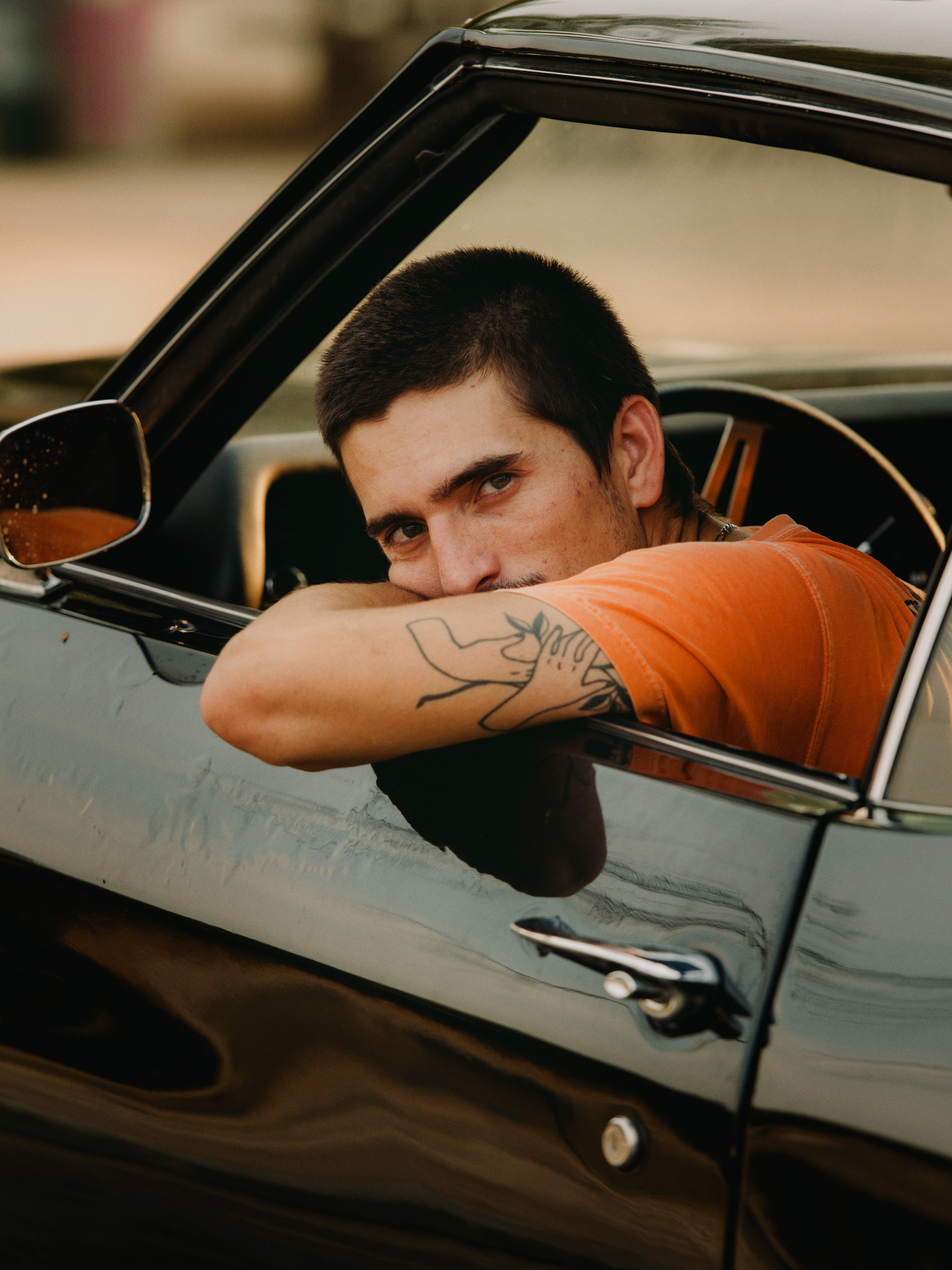Adult man in an orange t-shirt leaning out the driver's side window of a classic black car with a tattoo on his arm.
