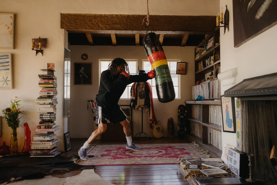 A person wearing boxing gloves practicing punching a rasta colored heavy bag inside a residential living room with book shelves.