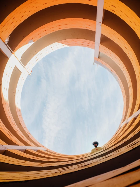 Low-angle shot of a Black person looking up through a large, orange, circular architectural structure against a bright blue sky.