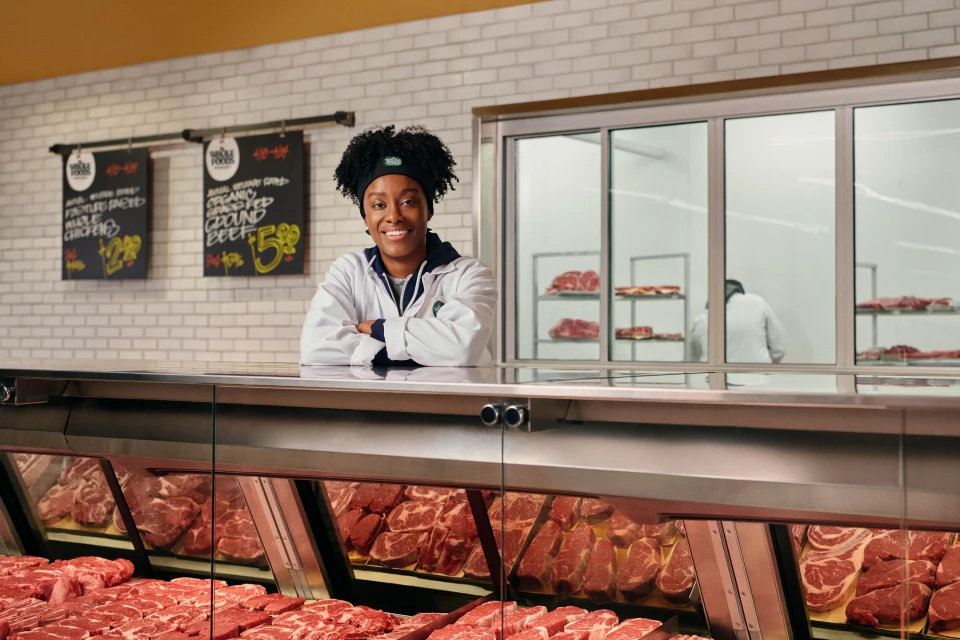 An African American woman smiles while working at a butcher counter filled with cuts of raw beef in a grocery store.