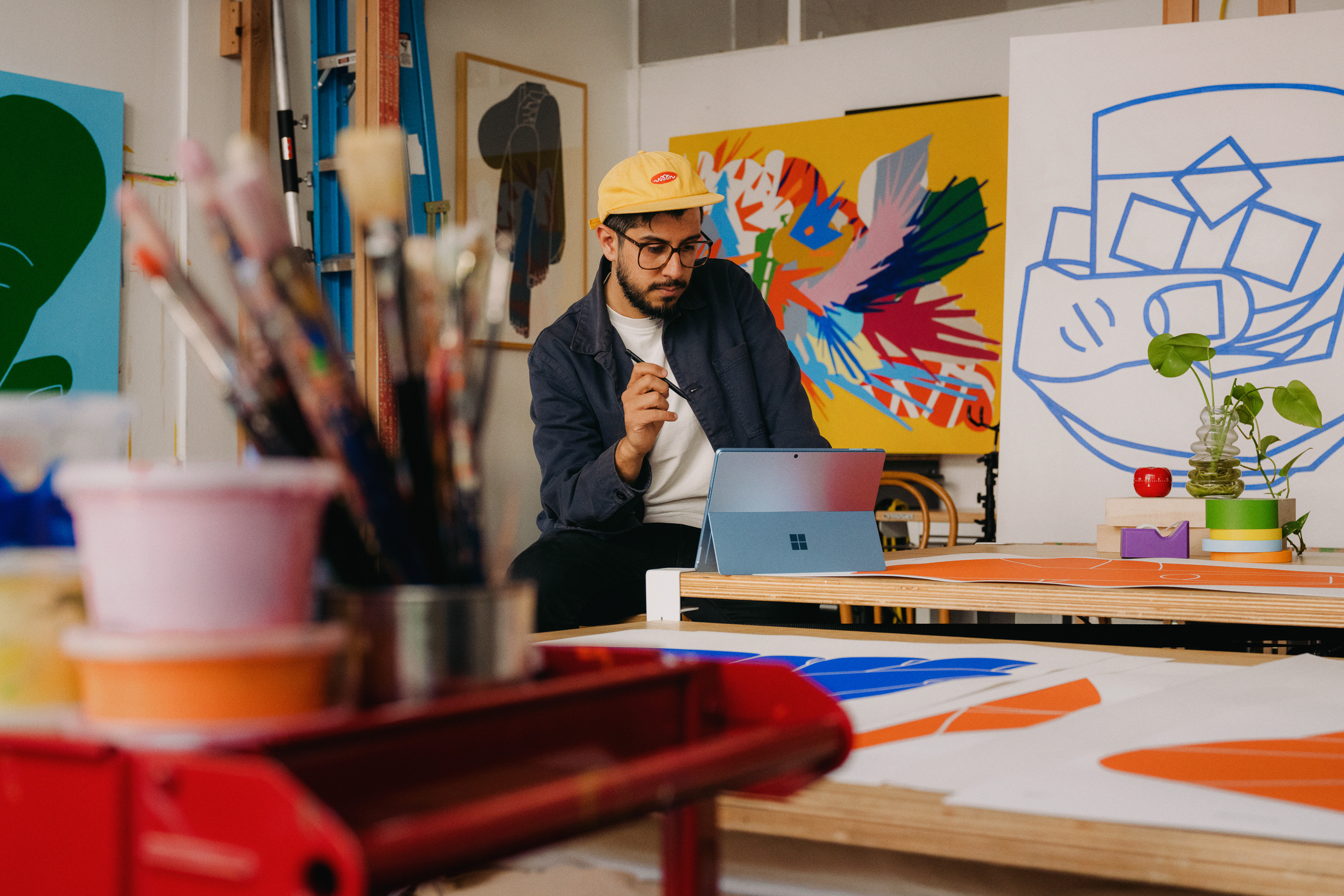 Male artist with glasses sitting in art studio at desk, using tablet with paint brushes, art supplies, and colorful paintings.