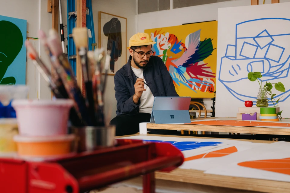 Male artist with glasses sitting in art studio at desk, using tablet with paint brushes, art supplies, and colorful paintings.