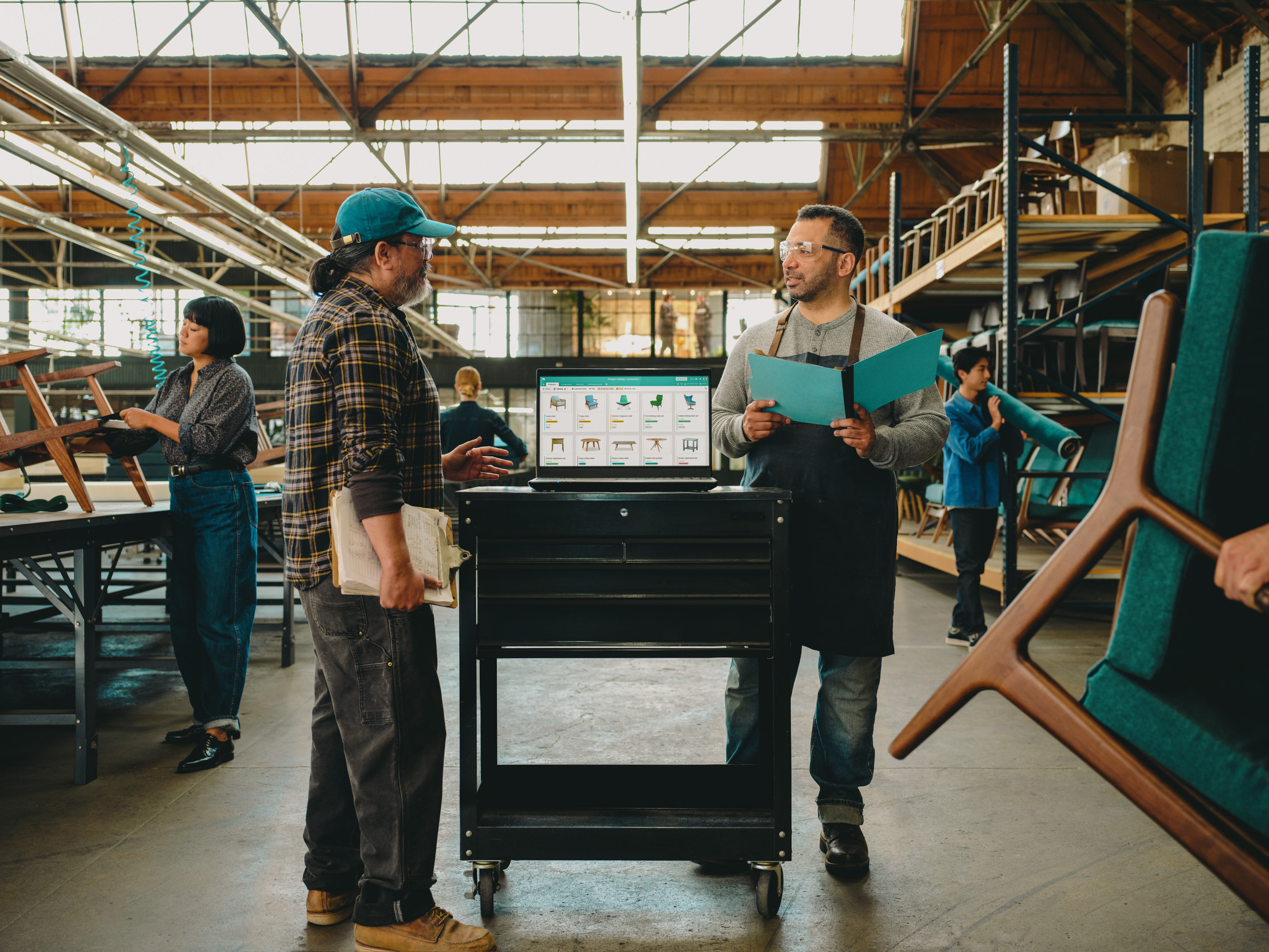 Two men in a furniture workshop examine a laptop displaying product images, with a woman working on a chair in the background.