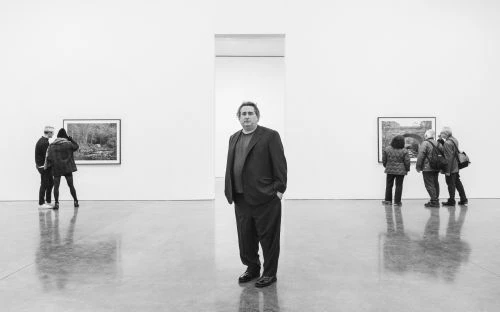 Monochrome shot of an adult male standing in an art gallery, with artwork and other gallery visitors in the background.