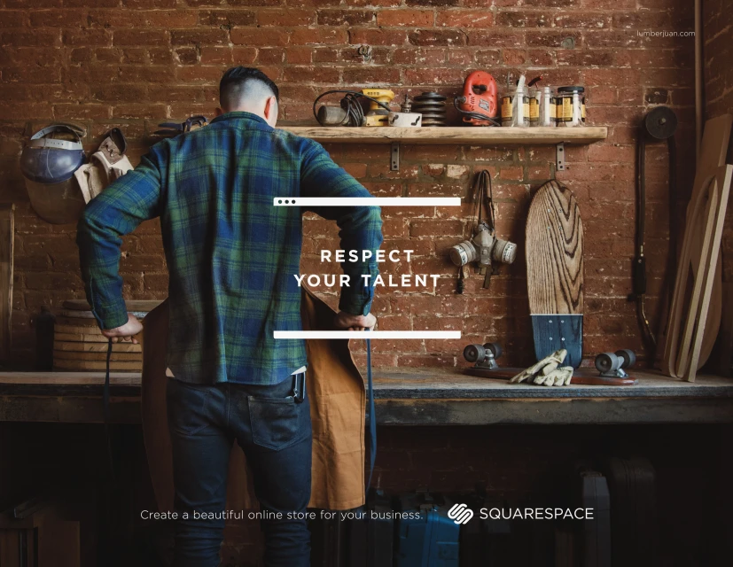 Carpenter in blue plaid shirt and dark jeans stands at a wooden workbench in his cluttered brick-walled shop. Craft tools on shelves.