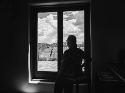 Person looks out a window at the landscape; clouds fill the sky, visible from indoors with natural light and monochrome tones.