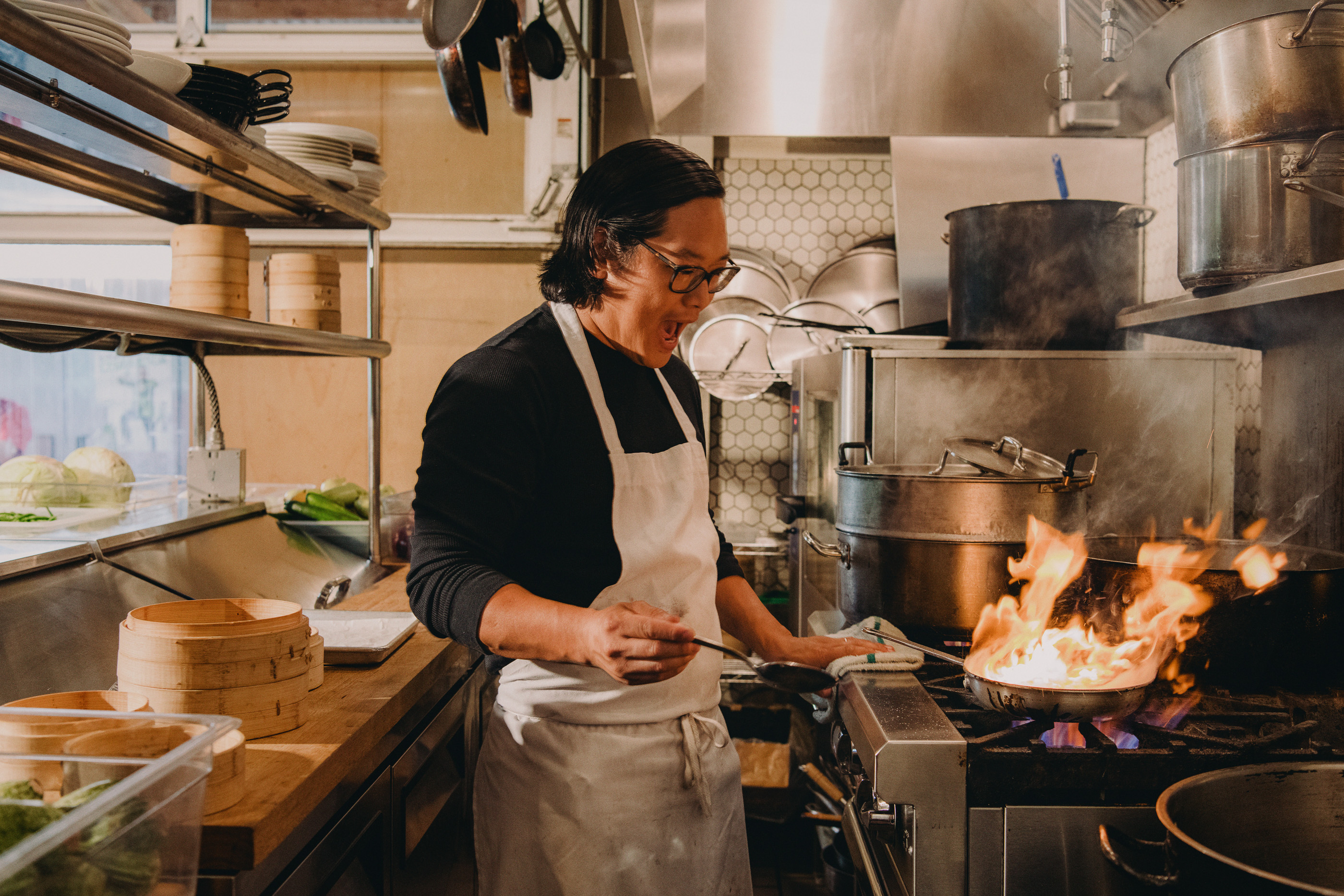 A chef with glasses cooks in a commercial kitchen with flames rising from the pan on the stove, surrounded by kitchenware.
