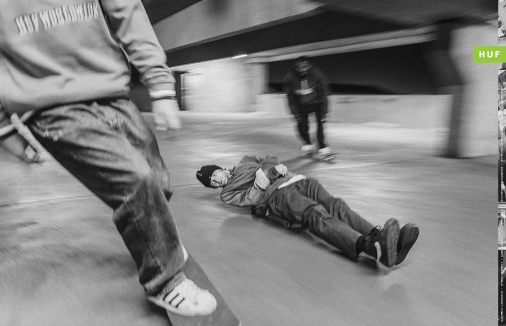 Black and white shot of skateboarders in motion, one lying down, in an indoor setting with the HUF logo visible, showcasing skateboarding action.